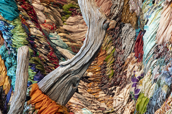 Two branches of weathered, grey driftwood are positioned vertically in the foreground in the left and center of the photo, against an undulating backdrop made of woven strips of colorful, patterned fabrics.