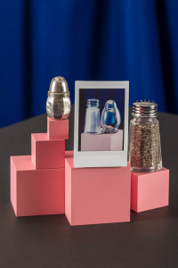 A still-life photograph featuring a small metal saltshaker on the left and a glass and metal peppershaker on the right, with a smaller photograph of a different salt and pepper shaker set in the middle. All of the objects are arranged on top of pink blocks of various sizes, set against a dark grey and blue background.